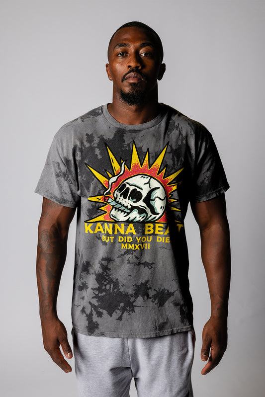 A young man with a shaved head stands against a light background wearing a gray tie-dye t-shirt. The shirt features a graphic design of a skull with spiky yellow rays around it, reminiscent of a sun. Below the skull is text that reads "KANNA BEAST BUT DID YOU DIE?" The man has a neutral expression and is looking directly at the camera.