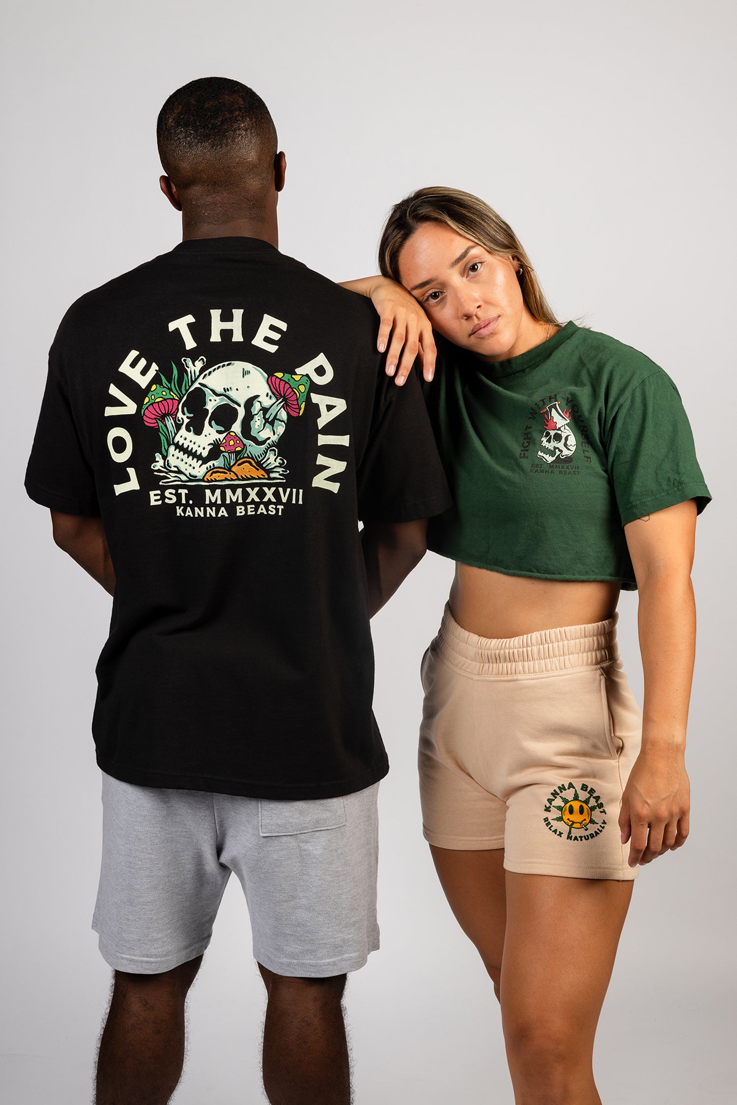 Two models posing together, the female model with her arm on the male model's shoulder wearing a green crop top and cream shorts. The male model is facing backwards with a black shirt and gray shorts.