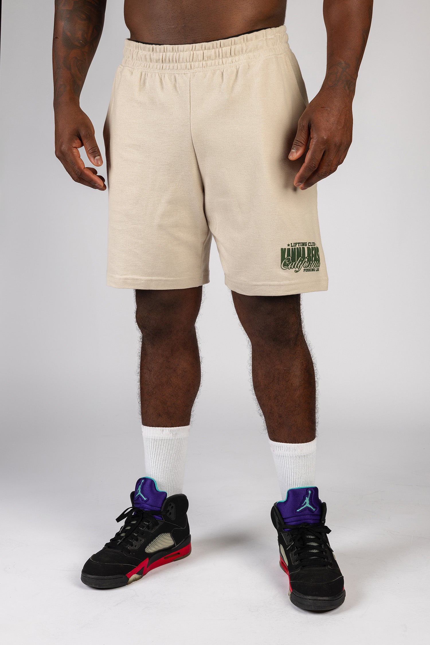 A male model wearing sand knee length shorts with the Kanna Beast brand and motto 'Lifting Club, Pushing LBs' in green lettering on the bottom right corner.
