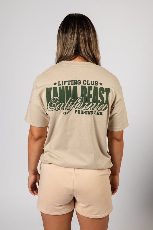 Female model showing the back of the sand Kanna Beast graphic t-shirt featuring Kanna Beast logo and motto 'Lifting Club, Pushing LBs' in green on.