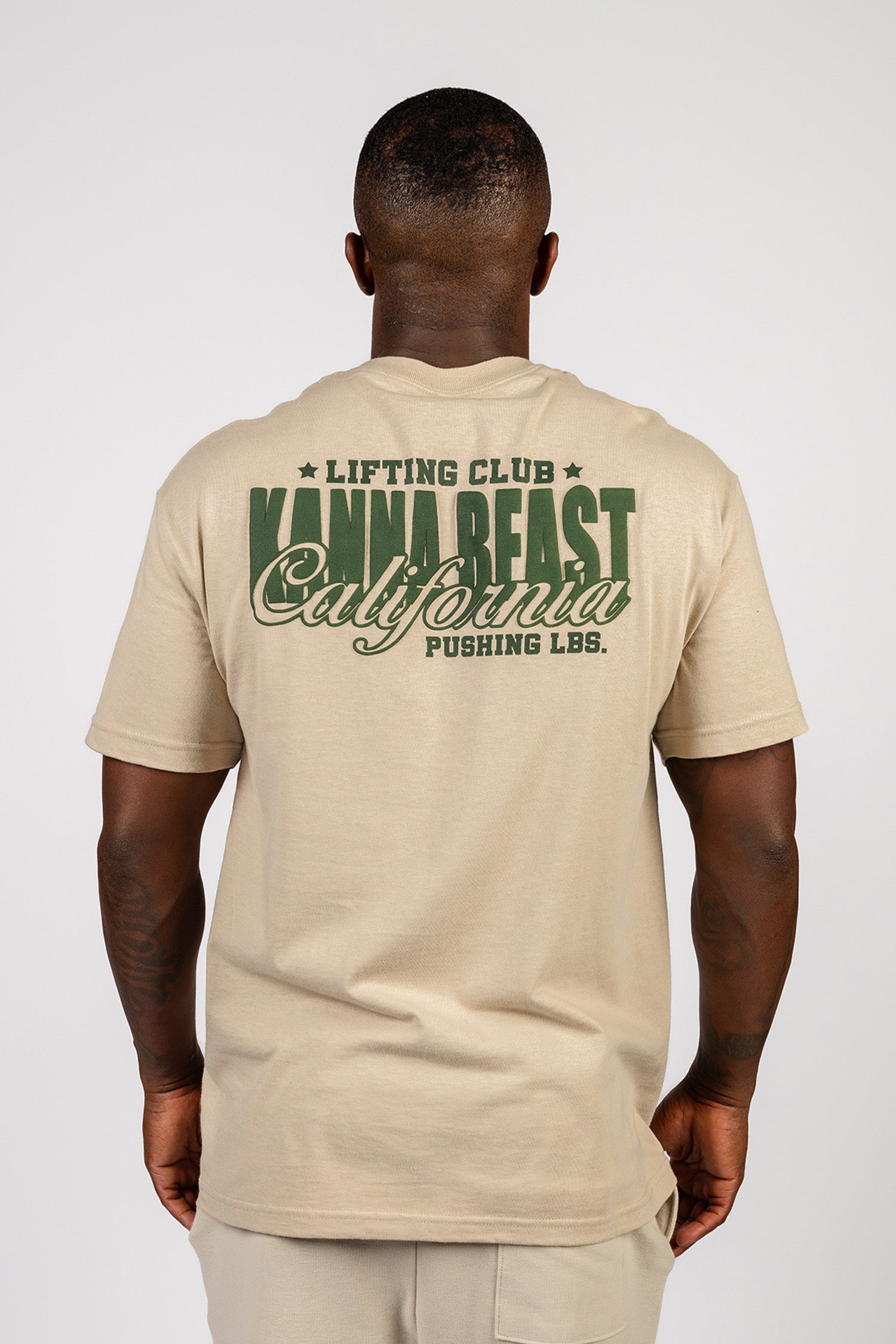 Male model showing the back of the sand Kanna Beast graphic t-shirt featuring Kanna Beast logo and motto 'Lifting Club, Pushing LBs' in green.