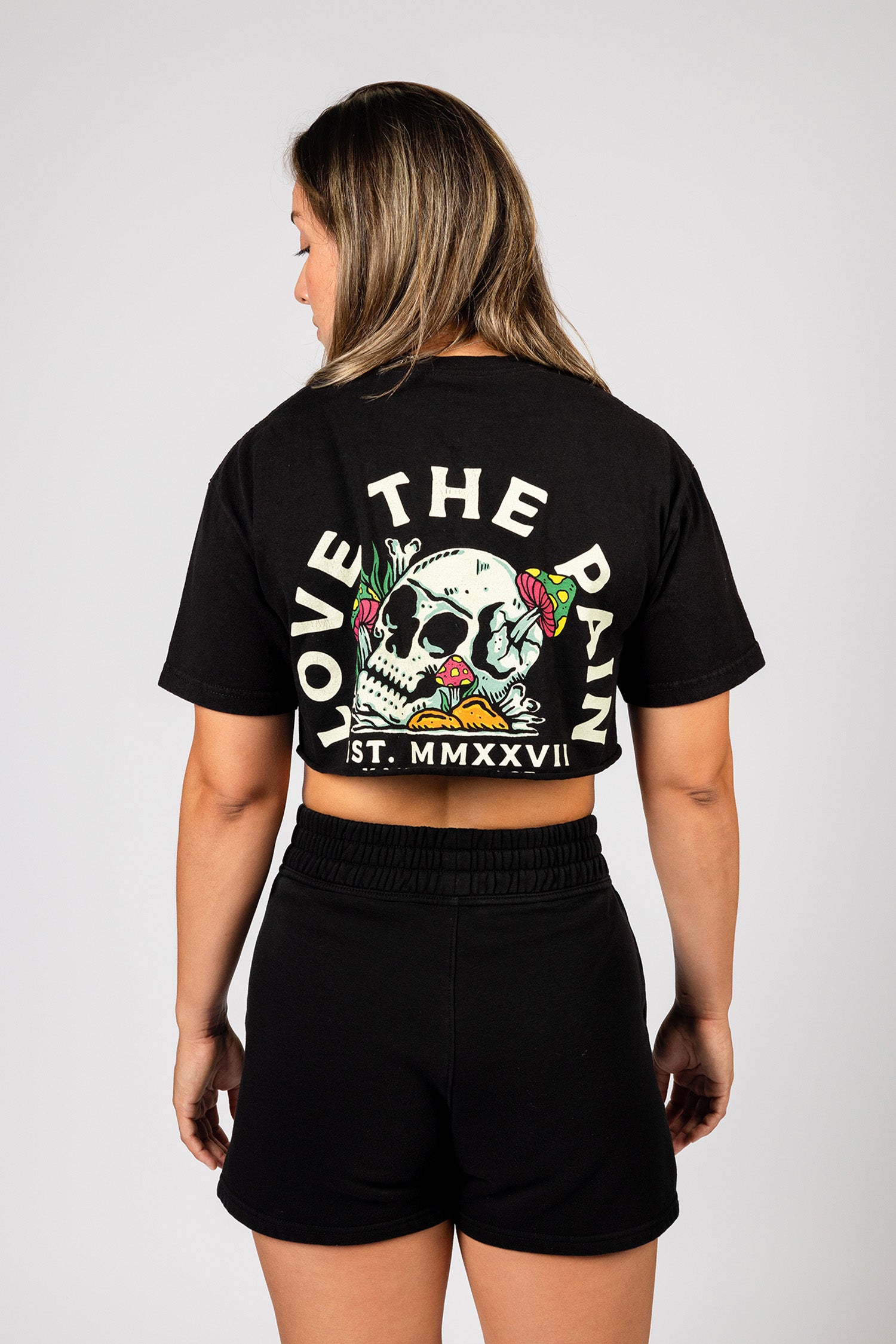 Showcasing the back of the shirt, a female model wears a cropped black Kanna Beast t-shirt with ‘Love The Pain’ graphic and skull design.