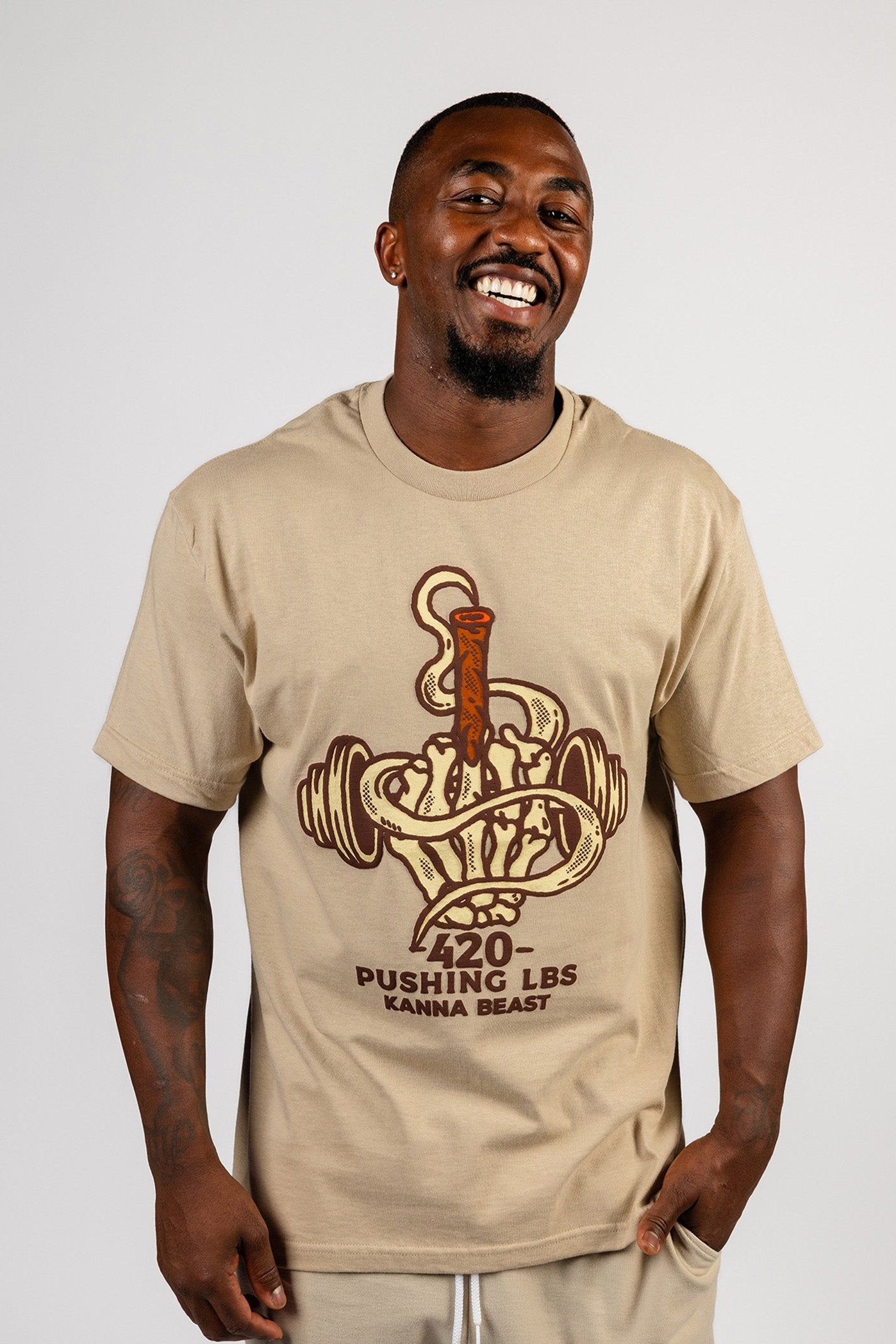 Male model with a large smile wearing beige Kanna Beast graphic t-shirt featuring a dumbbell and smoke design with the phrase ‘420 Pushing LBS'.