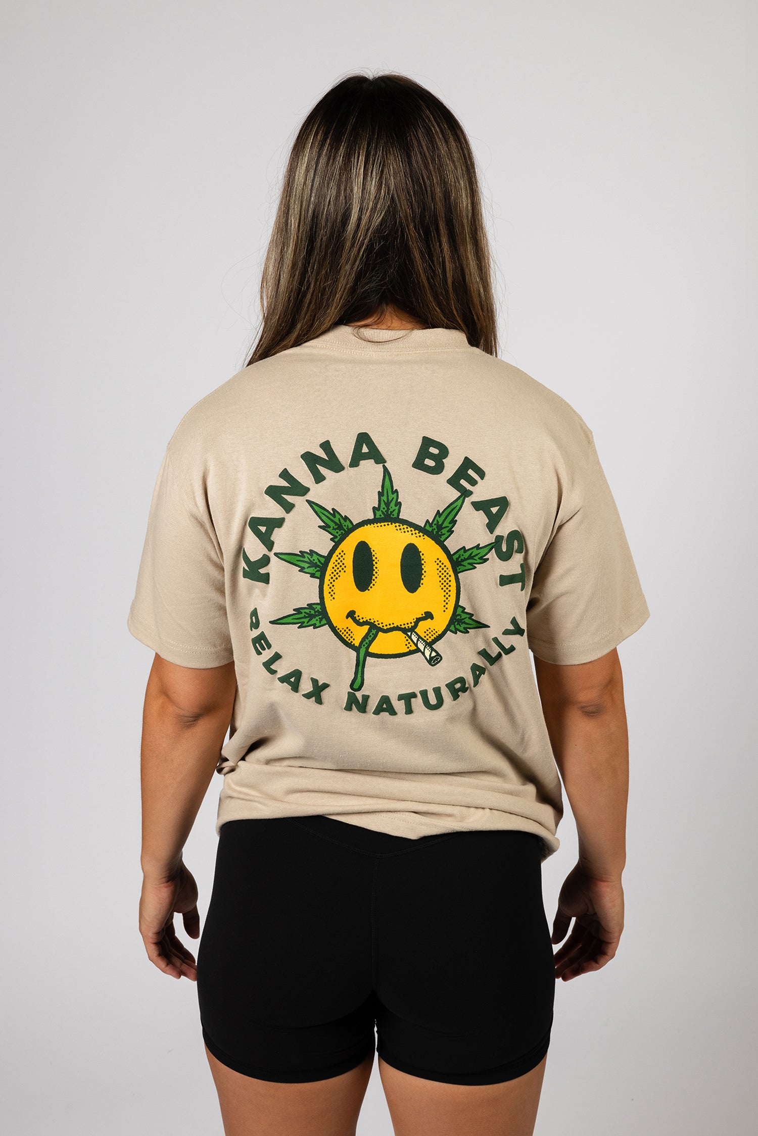 A young woman with long brown hair stands against a light background wearing a sand t-shirt. The shirt features a graphic design of a smiley face with spiky green leaves Surrounding the text is "Kanna Beast, Relax Naturally."
