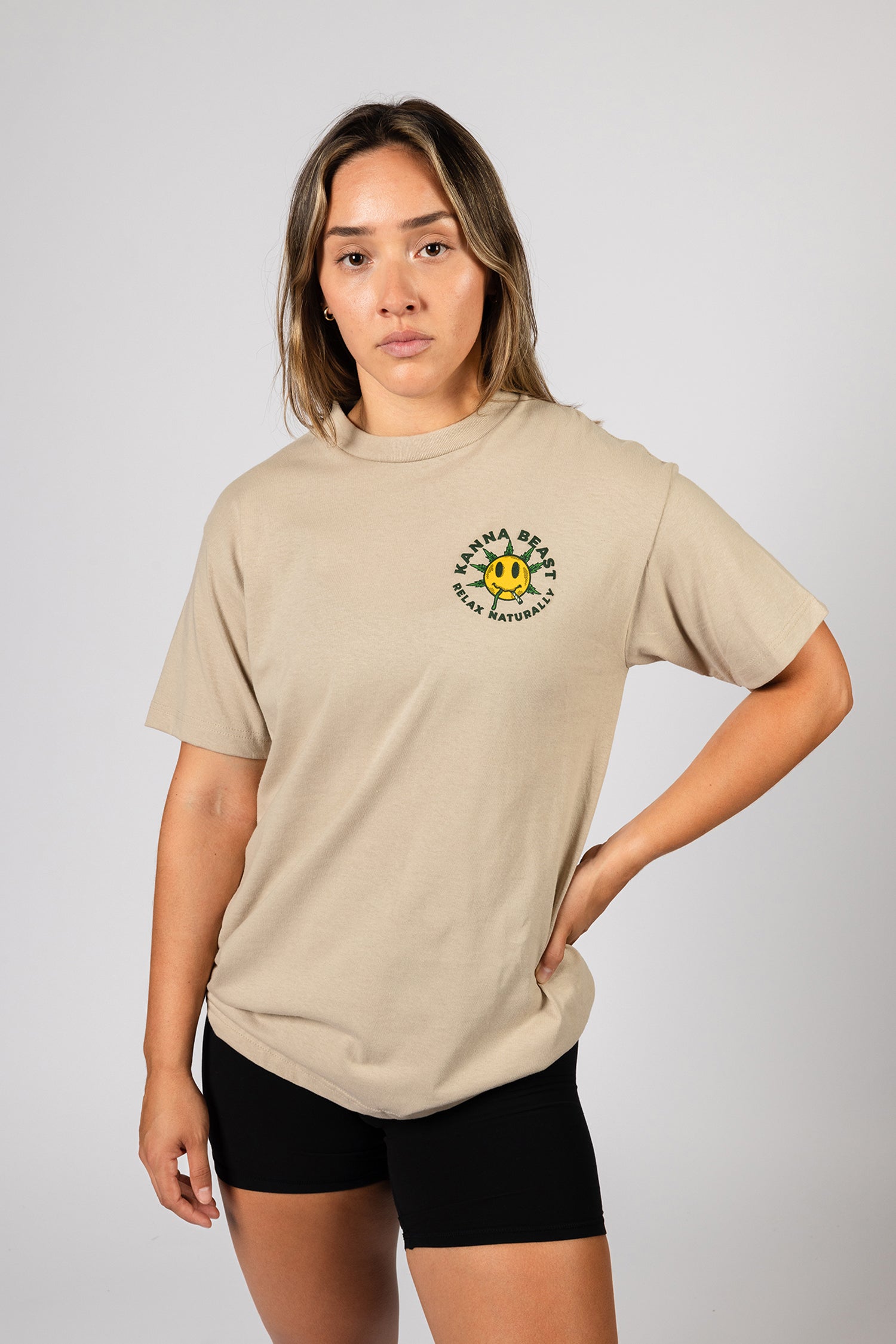A young woman with long brown hair stands against a light background wearing a sand t-shirt. The shirt features a graphic design of a smiley face with spiky green leaves Surrounding the text is "Kanna Beast, Relax Naturally." The woman has a neutral expression and is looking directly at the camera