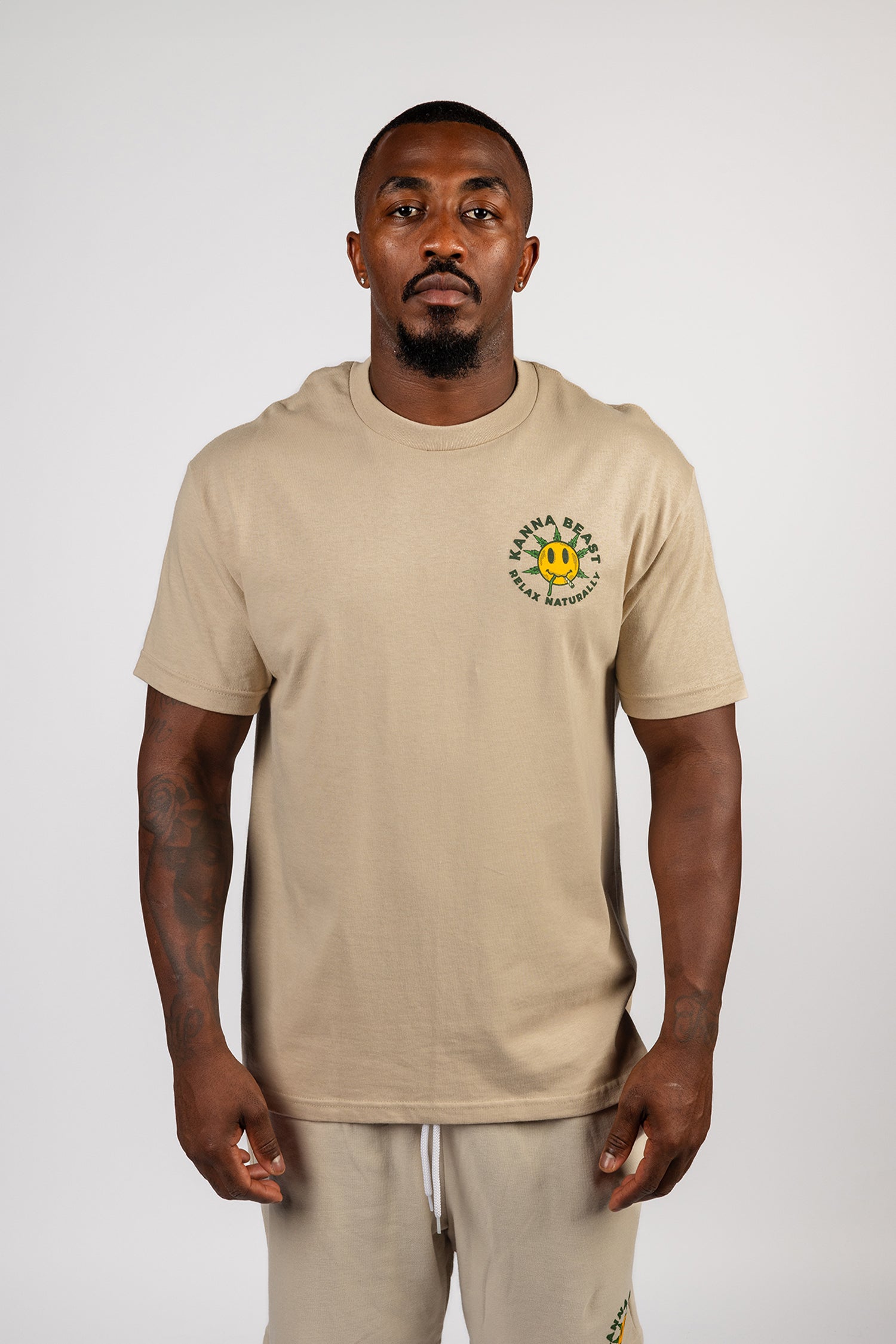 A young man stands against a light background wearing a sand t-shirt. The shirt features a graphic design of a smiley face with spiky green leaves Surrounding the text is "Kanna Beast, Relax Naturally." The man has a neutral expression and is looking directly at the camera