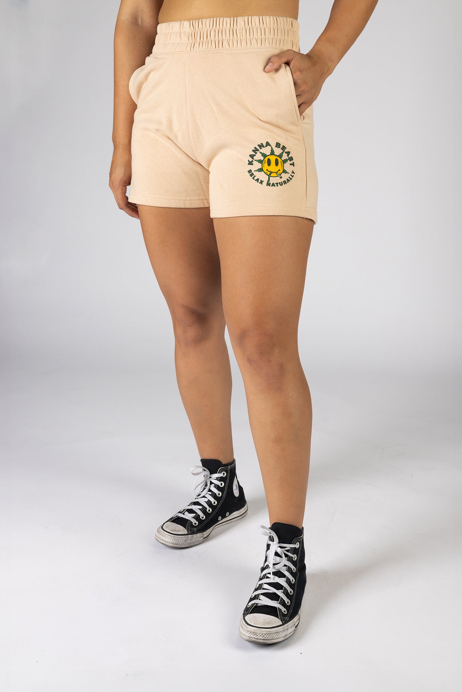 A woman posing in front of a light background wearing cream shorts The shorts feature a graphic design of a smiley face with spiky green leaves. Surrounding the graphic is "Kanna Beast, Relax Naturally."