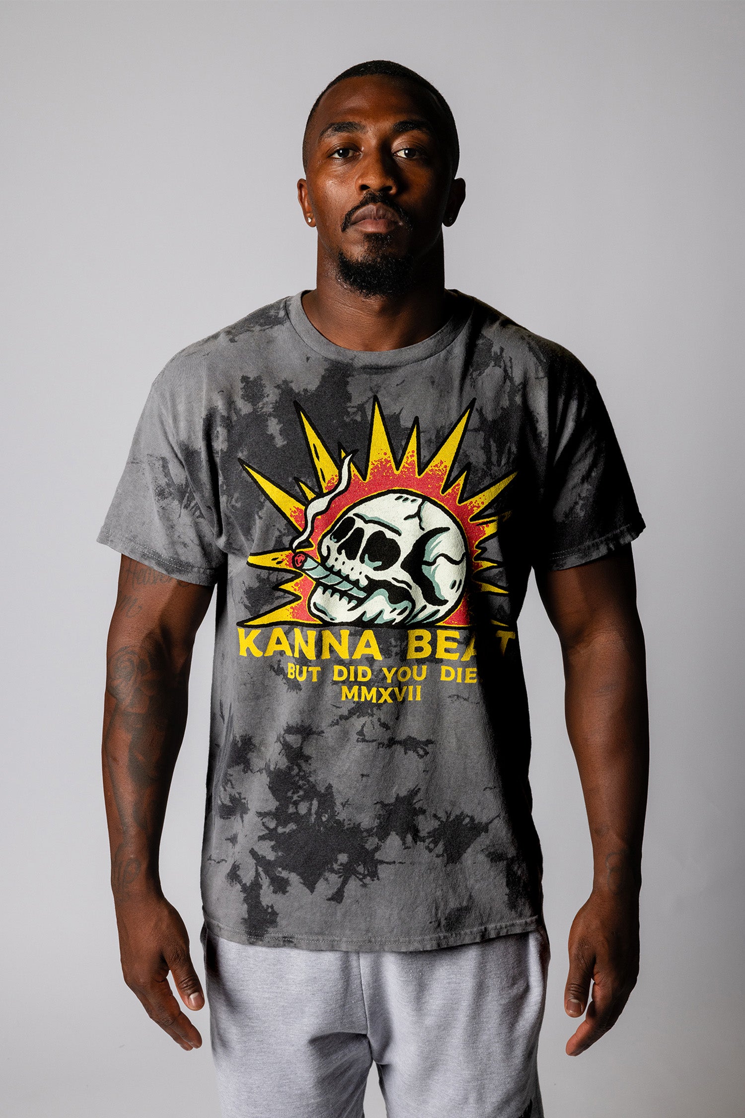 A young man with a shaved head  stands against a light background wearing a gray tie-dye t-shirt. The shirt features a graphic design of a skull with spiky yellow rays around it, reminiscent of a sun. Below the skull is text that reads "KANNA BEAST BUT DID YOU DIE?" The man has a neutral expression and is looking directly at the camera.