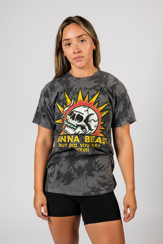 A young woman with long brown hair stands against a light background wearing a gray tie-dye t-shirt. The shirt features a graphic design of a skull with spiky yellow rays around it, reminiscent of a sun. Below the skull is text that reads "KANNA BEAST BUT DID YOU DIE?" The woman has a neutral expression and is looking directly at the camera.