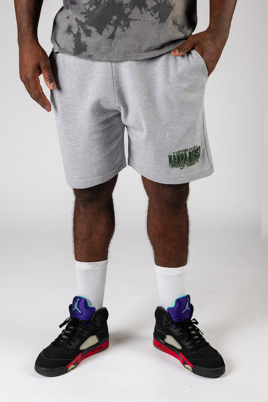 A male model wearing gray knee length shorts with hand in pocket,  the Kanna Beast brand and motto 'Lifting Club, Pushing LBs' in green lettering. 