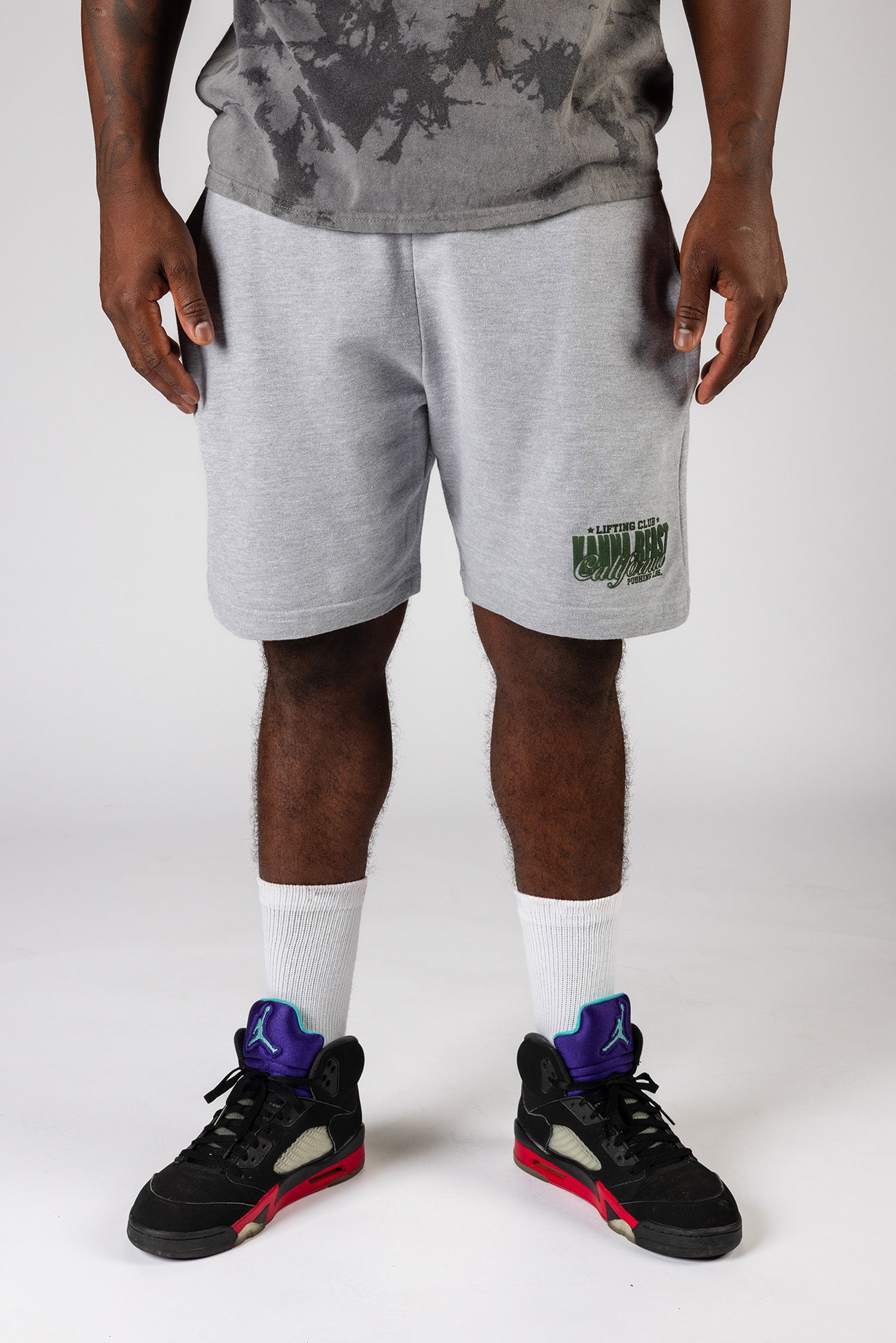 A male model wearing gray knee length shorts with the Kanna Beast brand and motto 'Lifting Club, Pushing LBs' in green lettering. 