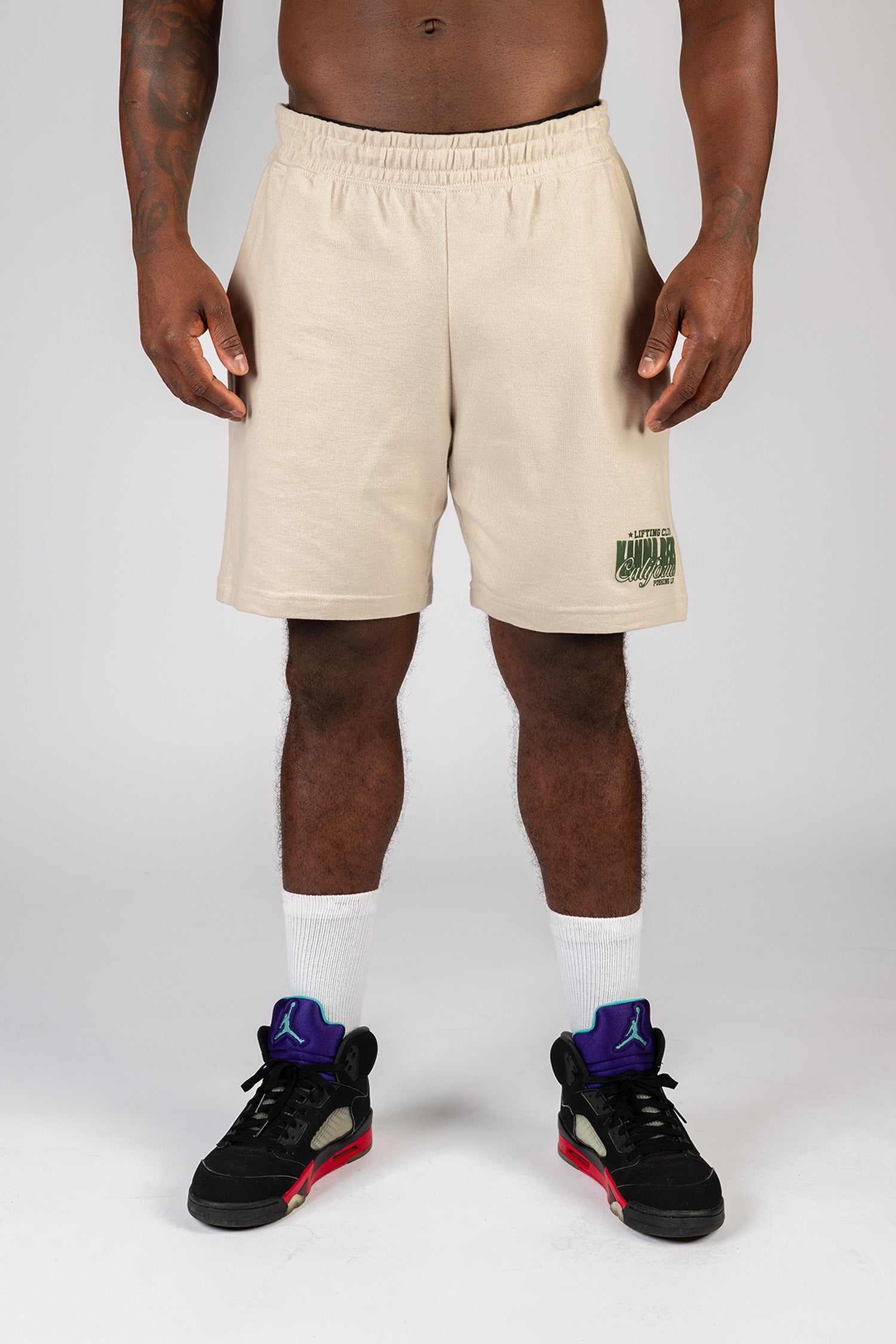 A male model wearing sand knee length shorts with the Kanna Beast brand and motto 'Lifting Club, Pushing LBs' in green lettering on the right corner. 