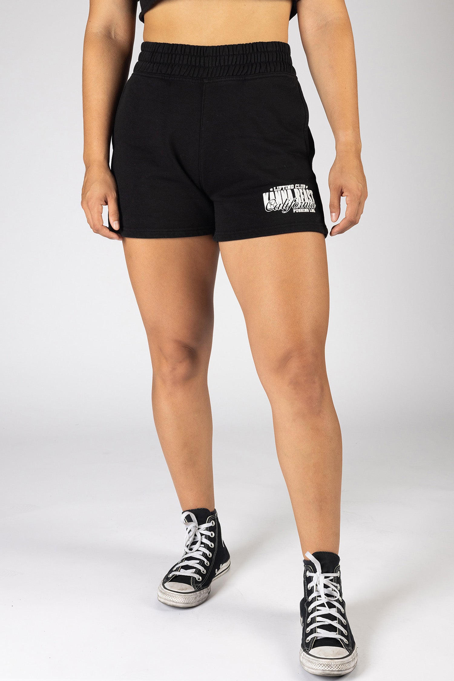 Female model wearing black Kanna Beast Lifting Club shorts. These shorts have motto 'Lifting Club, Pushing LBs' on left thigh in white.