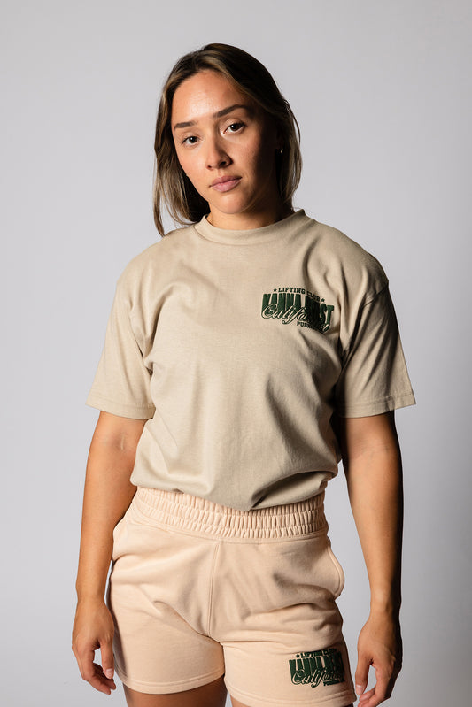 Female model wearing sand Kanna Beast graphic t-shirt featuring Kanna Beast logo and motto 'Lifting Club, Pushing LBs' in green on the left chest. 