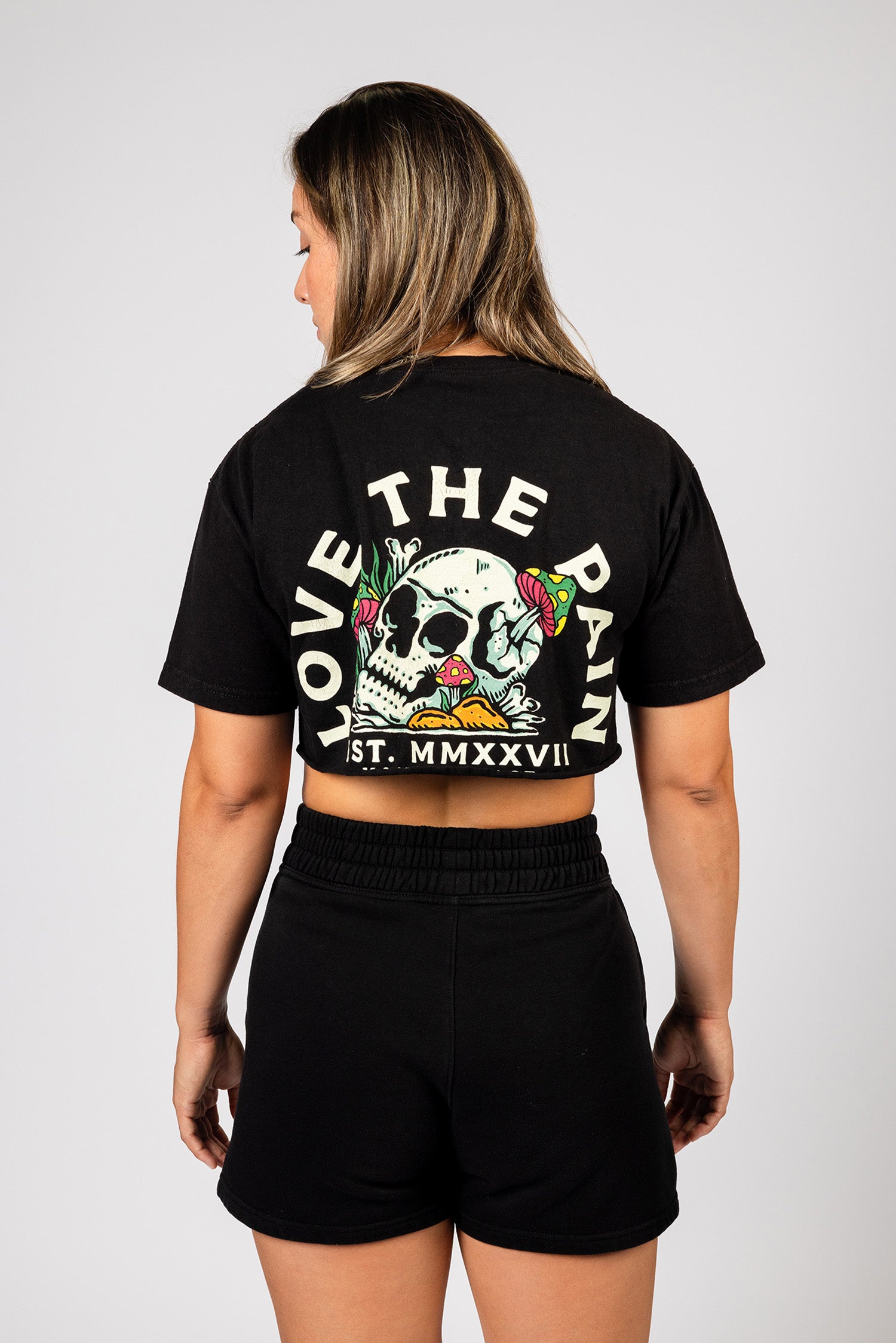 Showcasing the back of the shirt, a female model wears a cropped black Kanna Beast t-shirt with ‘Love The Pain’ graphic and skull design.
