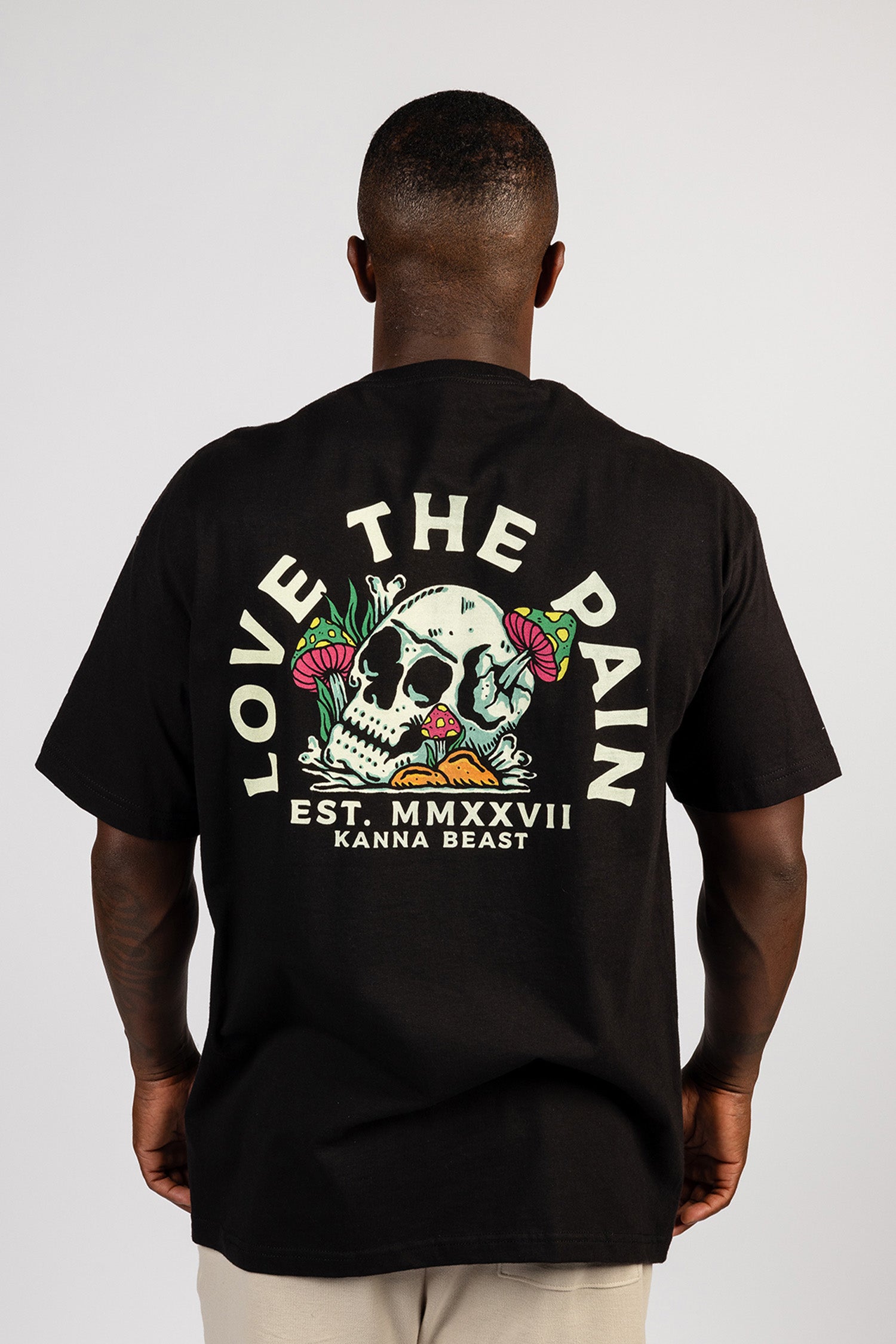 Showing the back of the shirt, a male model wears a black Kanna Beast t-shirt with ‘Love The Pain’ graphic and skull design.