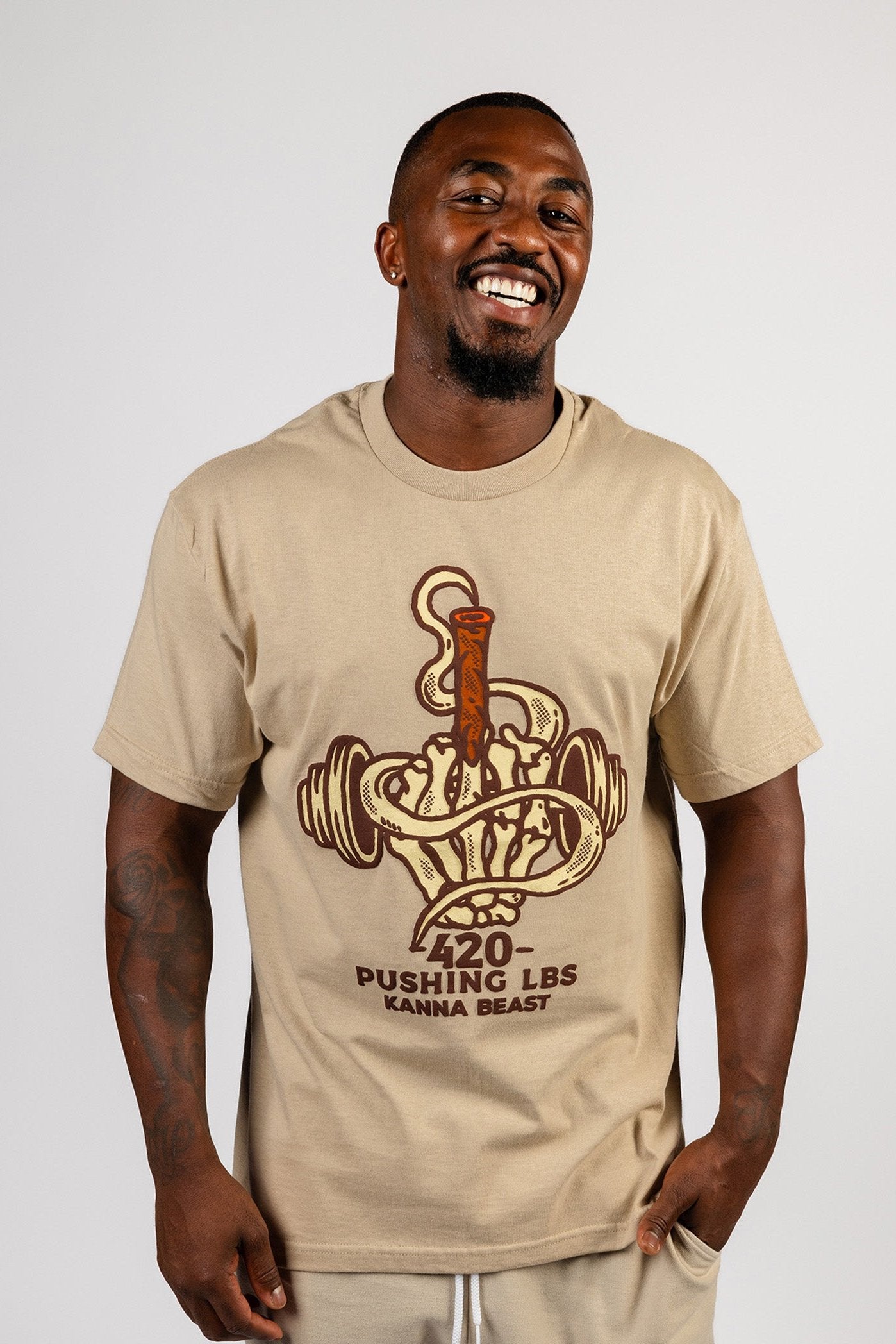 Male model with a large smile wearing beige Kanna Beast graphic t-shirt featuring a dumbbell and smoke design with the phrase ‘420 Pushing LBS'.