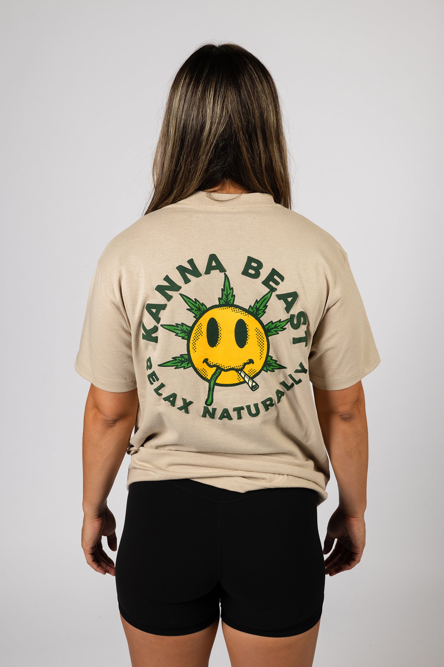 A young woman with long brown hair stands against a light background wearing a sand t-shirt. The shirt features a graphic design of a smiley face with spiky green leaves  Surrounding the text is "Kanna Beast, Relax Naturally."  