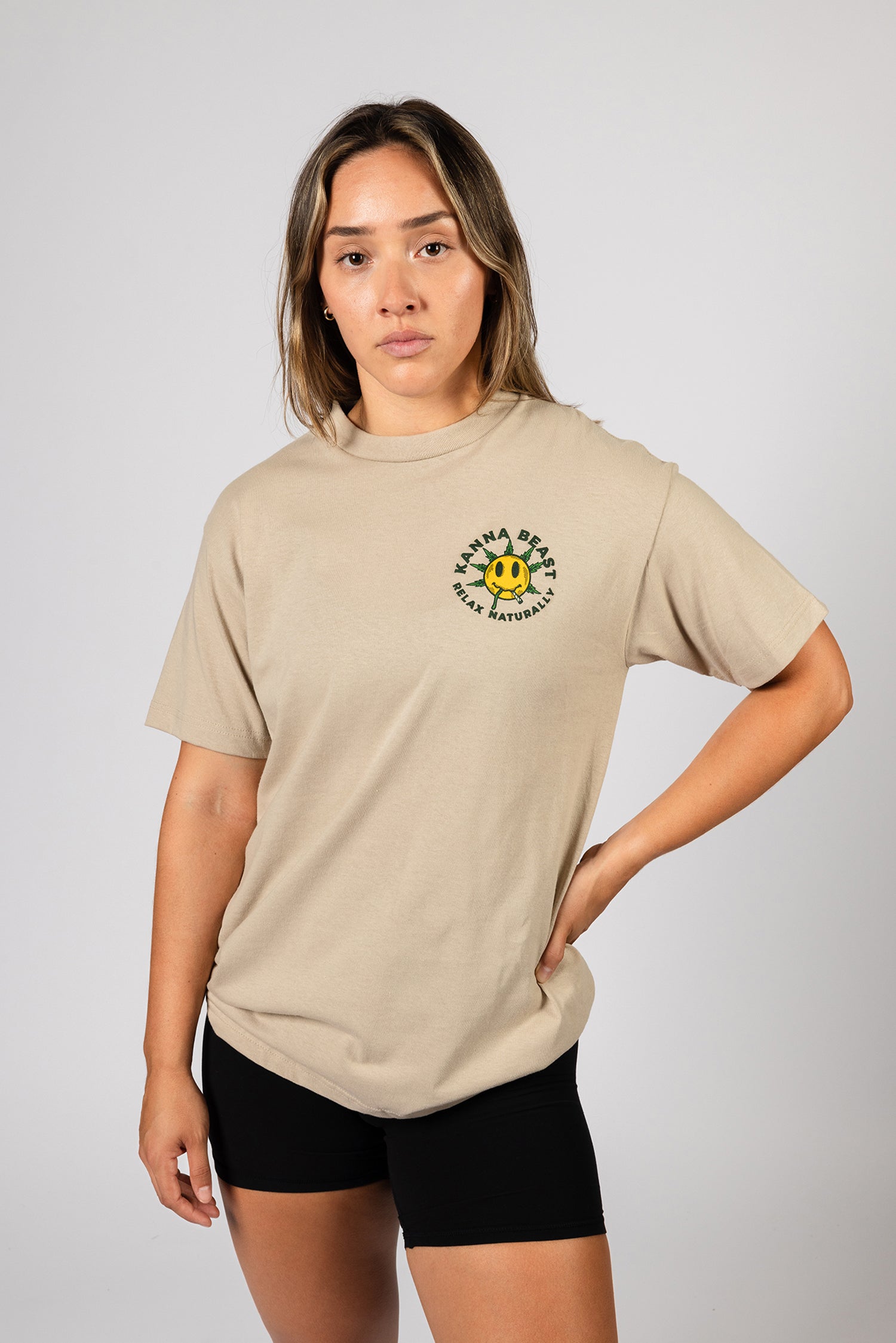 A young woman with long brown hair stands against a light background wearing a sand t-shirt. The shirt features a graphic design of a smiley face with spiky green leaves  Surrounding the text is "Kanna Beast, Relax Naturally."  The woman has a neutral expression and is looking directly at the camera