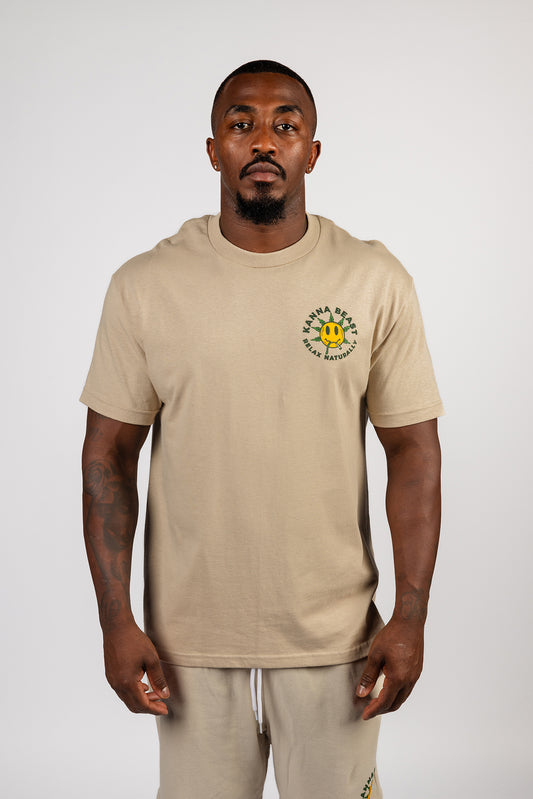 A young man stands against a light background wearing a sand t-shirt. The shirt features a graphic design of a smiley face with spiky green leaves  Surrounding the text is "Kanna Beast, Relax Naturally."  The man has a neutral expression and is looking directly at the camera