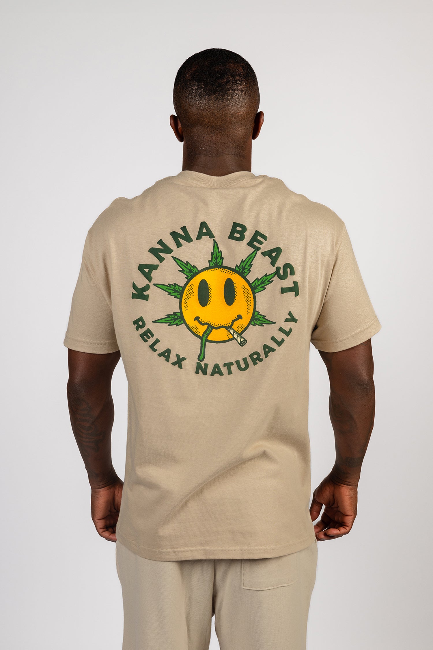 A young man stands against a light background wearing a sand t-shirt. The shirt features a graphic design of a smiley face with spiky green leaves  Surrounding the text is "Kanna Beast, Relax Naturally." 