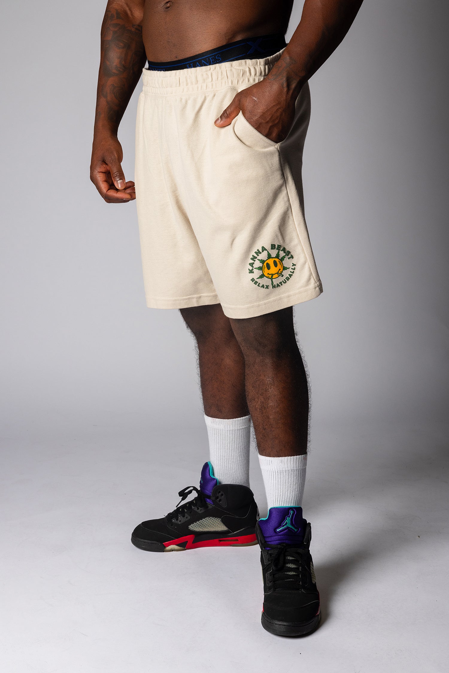 Man wearing sand athletic shorts with Kanna Beast Smiley Face graphic and text "Relax Naturally" paired with white socks and black sneakers