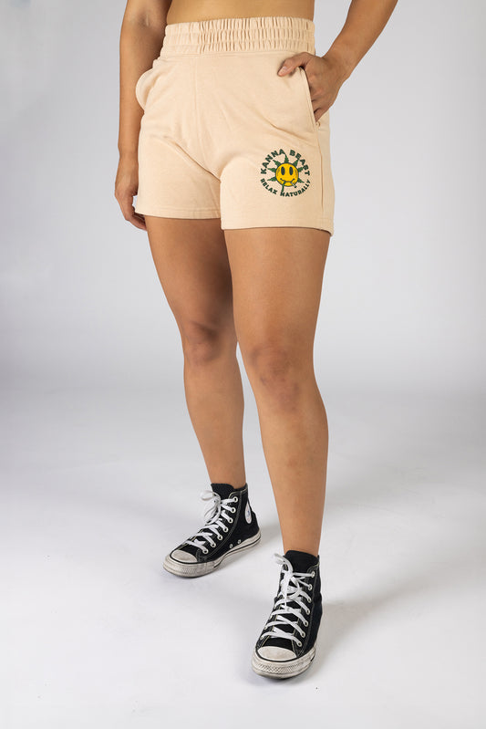 A woman posing in front of a light background wearing cream shorts The shorts feature a graphic design of a smiley face with spiky green leaves.  Surrounding the graphic is "Kanna Beast, Relax Naturally." 